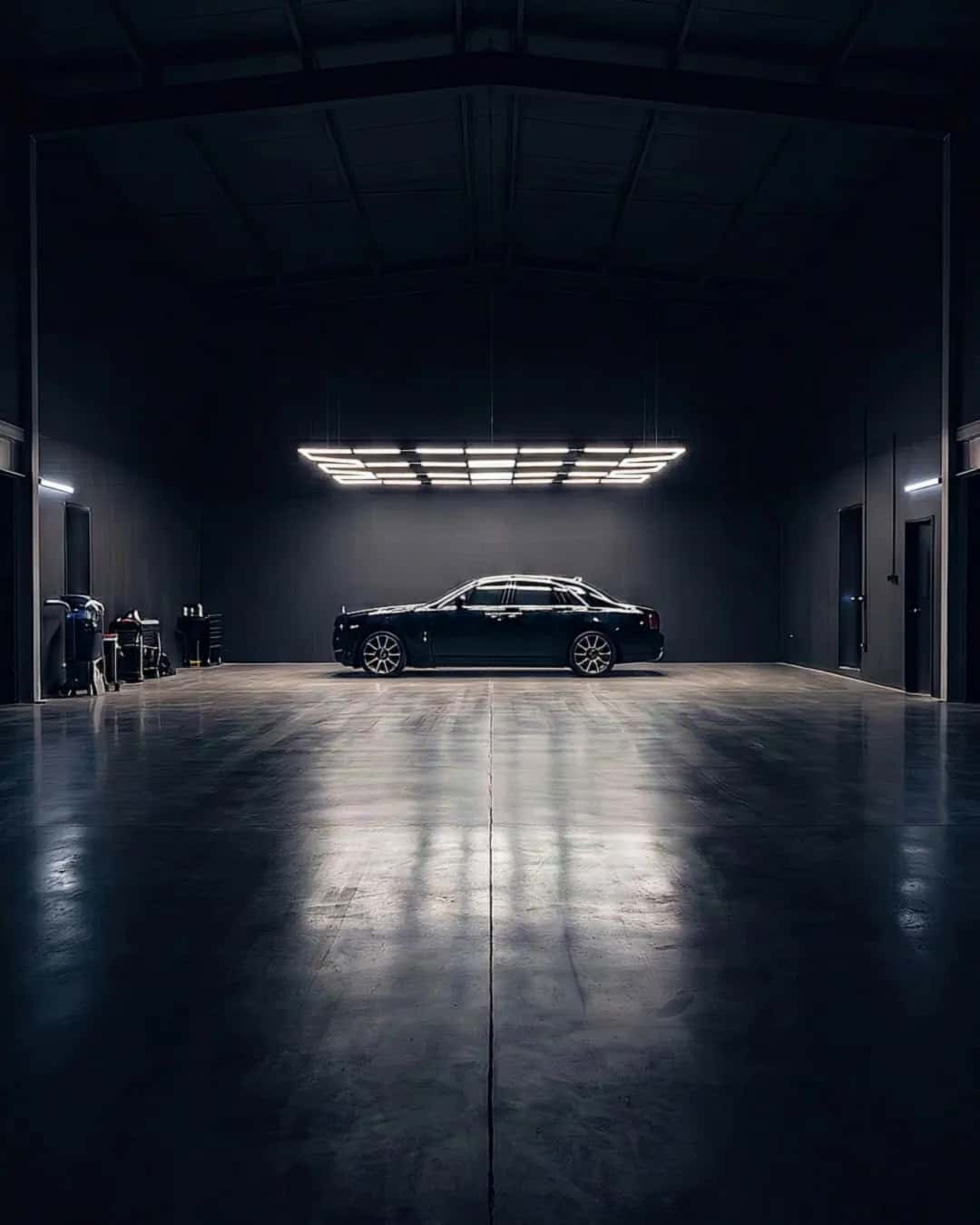 A dark, empty detailing bay at night seen down its centre line: one dark saloon parked at the far end under a cool white ceiling light, whose reflection runs across the polished floor.
