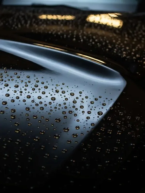 Close-up of round water beads standing on a dark glossy body panel, a pale reflection curving across it and two warm lights mirrored in the dark background.