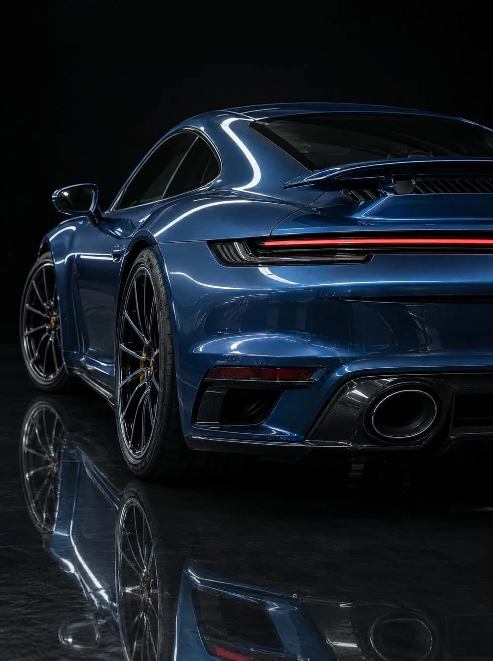 The glossy rear three-quarter of a deep blue Porsche 911 Turbo S, a curved highlight running the length of its roofline and its reflection in the dark floor.