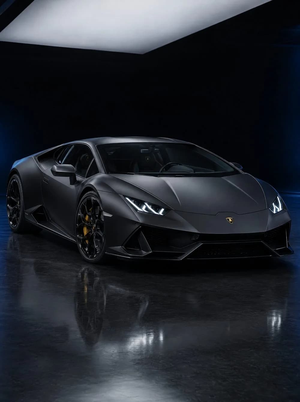 A satin black Lamborghini Huracán seen from the front three-quarter on a dark reflective floor, lit by one broad soft panel overhead with a faint blue glow behind it.