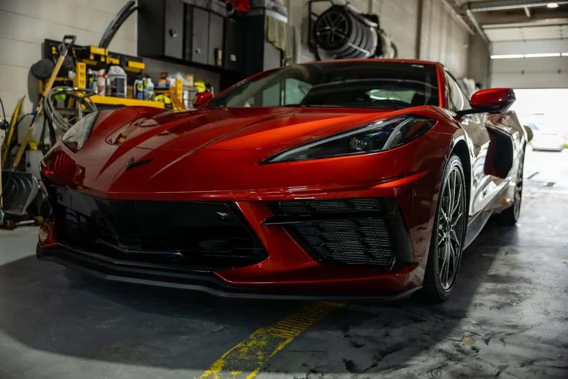 A red Chevrolet Corvette parked nose-out in a working shop bay, a wall of tools and bottles behind it, wheels stacked overhead and daylight through the open bay door.