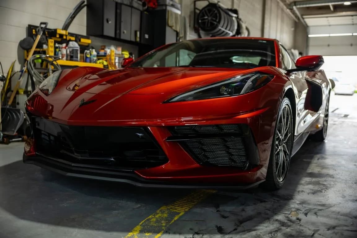 A red Chevrolet Corvette parked nose-out in a working shop bay, a wall of tools and bottles behind it, wheels stacked overhead and daylight through the open bay door.