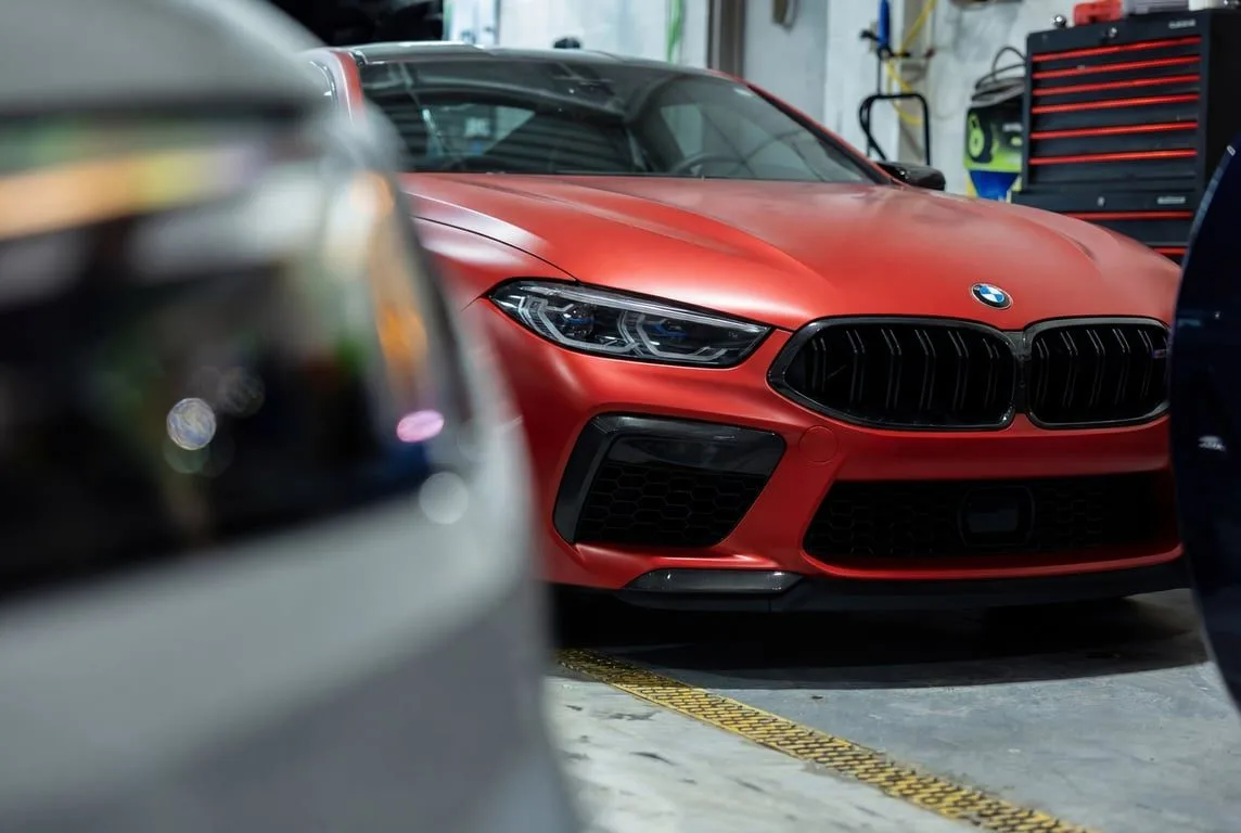 A satin red BMW M8 parked in a shop, seen past the blurred rear of another car in the foreground.